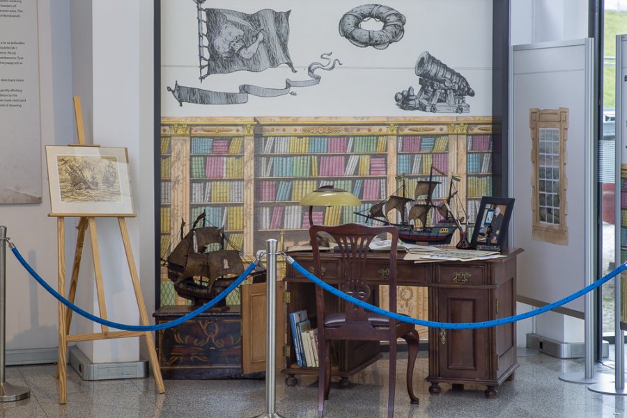 Fragment wystawy  „Akwarela, pastel, piórko – w służbie mistrza ilustracji Eugeniusza Koczorowskiego”: drewniane biurko, na którym stoją: lampa, model galeonu i zdjęcie kmdr. Eugeniusza Koczorowskiego. Całość na tle ścianek, na których namalowano regał z książkami i okno // fot. materiały MMW