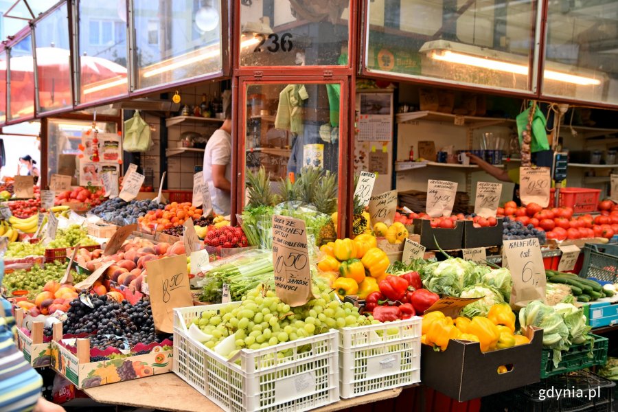 Stoisko z różnego rodzaju  owocami i warzywami, przy których umieszczono kartki z cenami i nazwami produktów. W tle widać sprzedawców, wiszące kolorowe reklamówki oraz półki ze słoikami i naczynami // fot. Sławomir Okoń