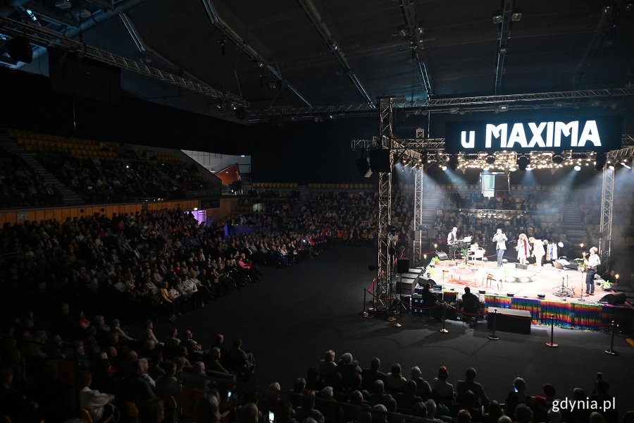Hala Polsat Plus Arena Gdynia, scena, dekoracja ze stołem i krzesłami, aktorzy na scenie, publiczność dookoła, nad sceną wyświetlacz z napisem "U Maxima"