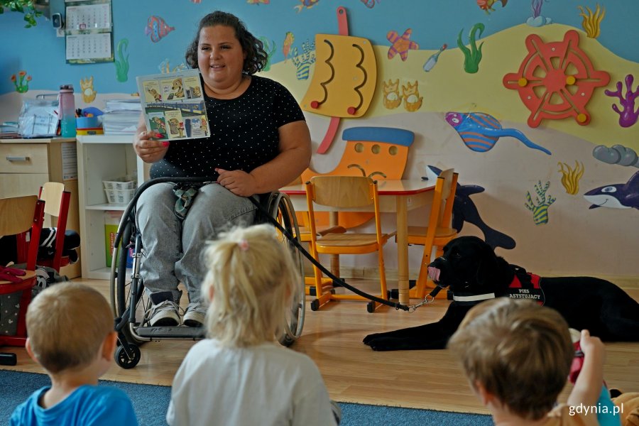 Marta Czachor z Wydziału Dostępności Urzędu Miasta Gdyni wraz z Delicją podczas warsztatów w Przedszkolu nr 60. Trzyma w ręku książeczkę dla dzieci z rysunkami na temat psa asystującego 