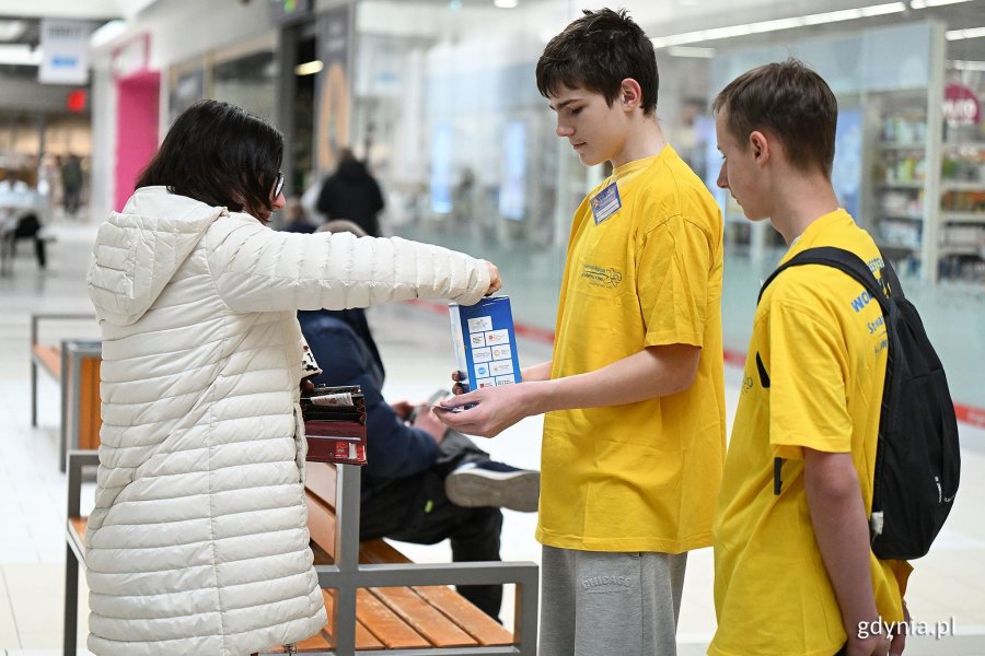 Kobieta w białej kurtce wrzuca pieniądze do skarbonki dw&oacute;ch młodych wolontariuszy w ż&oacute;łtych koszulkach