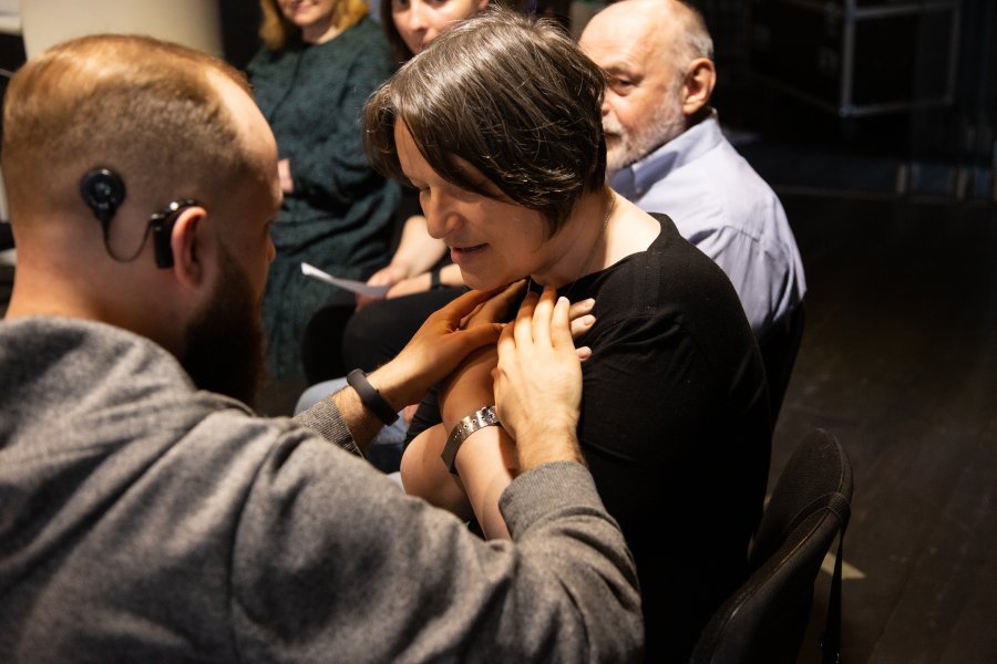 Na pierwszym planie mężczyzna i kobieta. Mężczyzna przykłada dłonie do klatki piersiowej. W tle inni uczestnicy warsztatów