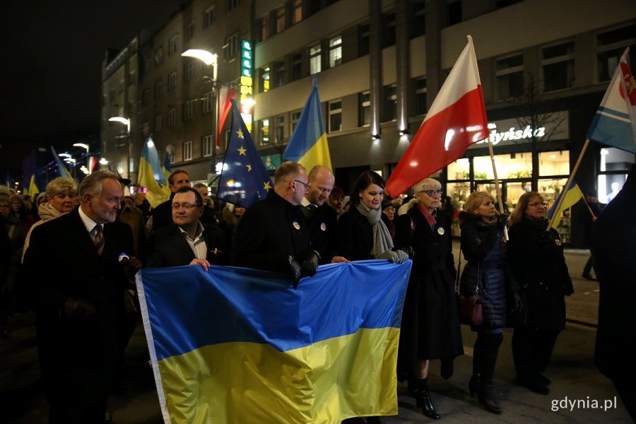 Tłum ludzi idzie wieczorem w marszu pokojowym ulicą Gdyni. W pierwszym rzędzie niosą niebiesko-żółtą flagę Ukrainy Wojciech Szczurek, prezydent Gdyni; Oleksandr Plodystyi, Konsul Generalny Ukrainy; wiceprezydenci Gdyni: Michał Guć, Bartosz Bartoszewicz, Katarzyna Gruszecka-Spychała; przewodnicząca Rady Miasta Gdyni - Joanna Zielińska.