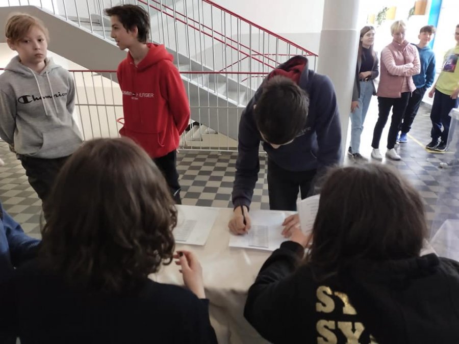 Na szkolnym korytarzu dwóch uczniów siedzi tyłem do obiektywu przy stole, jeden pochyla się nad stołem, w tle kolejne osoby czekają na swoją kolej. Widać fragment klatki schodowej.
