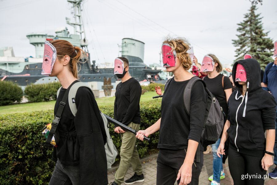 Uczestnicy akcji "Stop przemocy wobec narodu Białorusi" w maskach Łukaszenki. Są ubarni na czarno. Jedna z osób trzyma w ręce czarna pałkę. W tle okręt ORP Błyskawica // fot. Karol Stańczak
