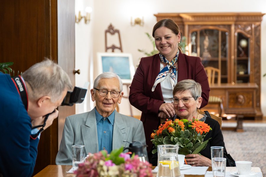 Prezydent Gdyni Aleksandra Kosiorek stojąca za plecami, siedzącej przy stole pary małżeńskiej