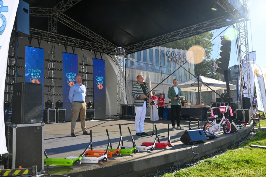 Trzy osoby na scenie, z brzegu sceny hulajnogi i nagłośnienie w tle niebieskie ścianki z logo festiwalu