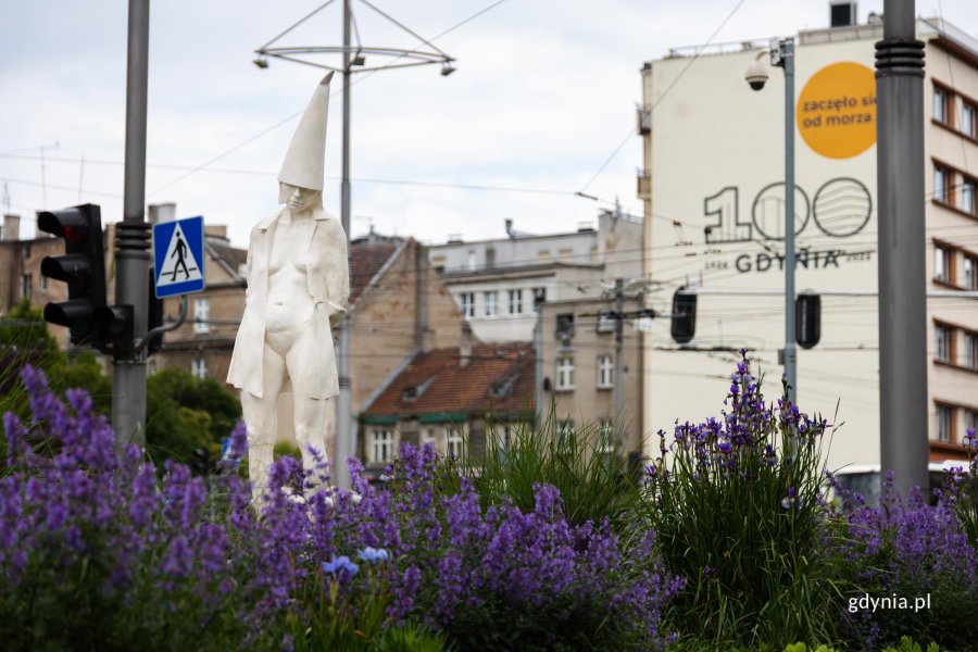 Rzeźba przedstawiająca kobietę z tr&oacute;jkątną czapką na głowie, stoi pomiędzy zielenią, w tle budynek z muralem "100 Gdynia. Zaczeło się od morza"