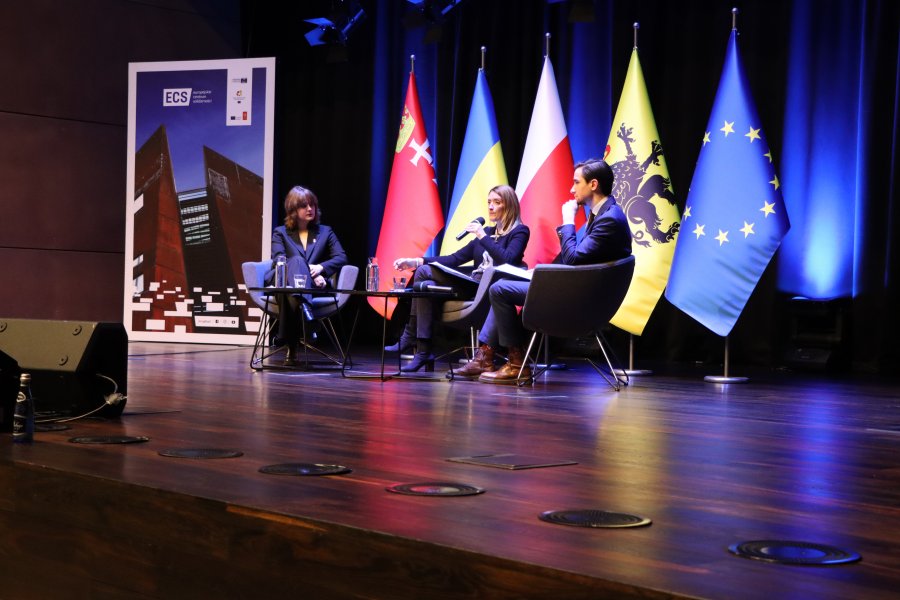 Trzy osoby siedzące na fotelach na scenie, w 5 flag: Unii Europejskiej, województwa Pomorskiego, Polski, Ukrainy, Gdańska.
