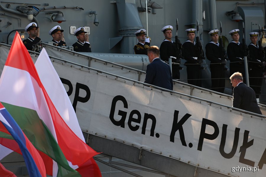 Ministrowie obrony państw Grupy Wyszehradzkiej wchodzą na pokład okrętu ORP "Gen. K. Pułaski". Na pierwszym planie powiewają flagi Polski, Węgier, Słowacji i Czech. Na okręcie w szeregu i na baczność stoją żołnierze Marynarki Wojennej // fot. Michał Puszczewicz