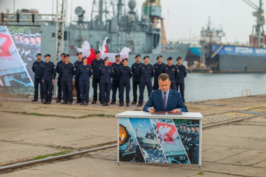 Minister obrony narodowej Mariusz Błaszczak podczas podpisania umowy na zakup nowych okrętów dla Marynarki Wojennej. W tle stojący na nabrzeżu marynarze i zacumowane okręty // fot.  bsmt Michał Pietrzak 