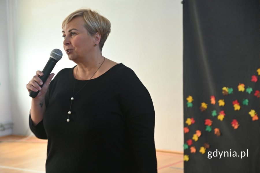 Stojąca kobieta mówiąca do mikrofonu w szkolnej hali sportowej
