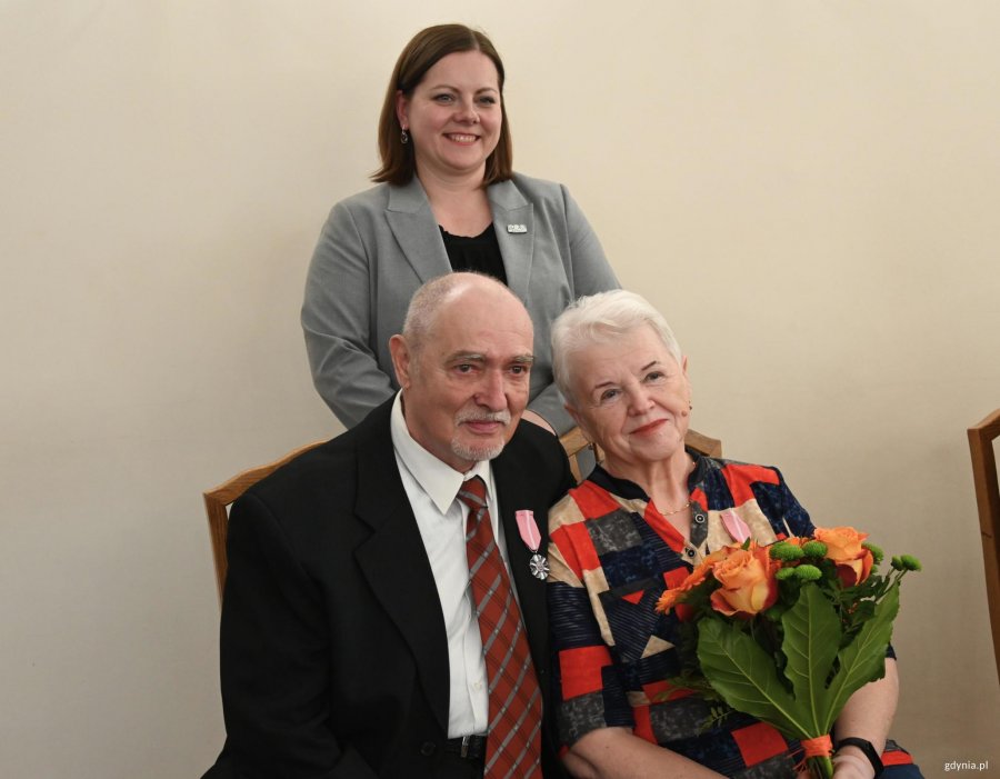 Para starszych ludzi na krzesłach. Za nimi stoi kobieta w szarej marynarce
