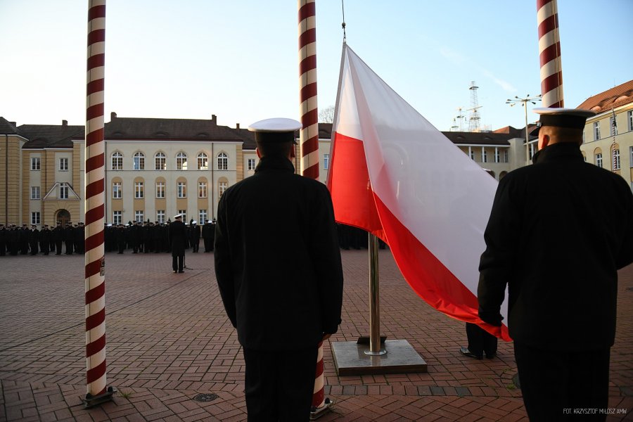 Dwaj mężczyźni w mundurach marynarskich stoją tyłem do fotografa, na maszt wciągana jest flaga Polski.