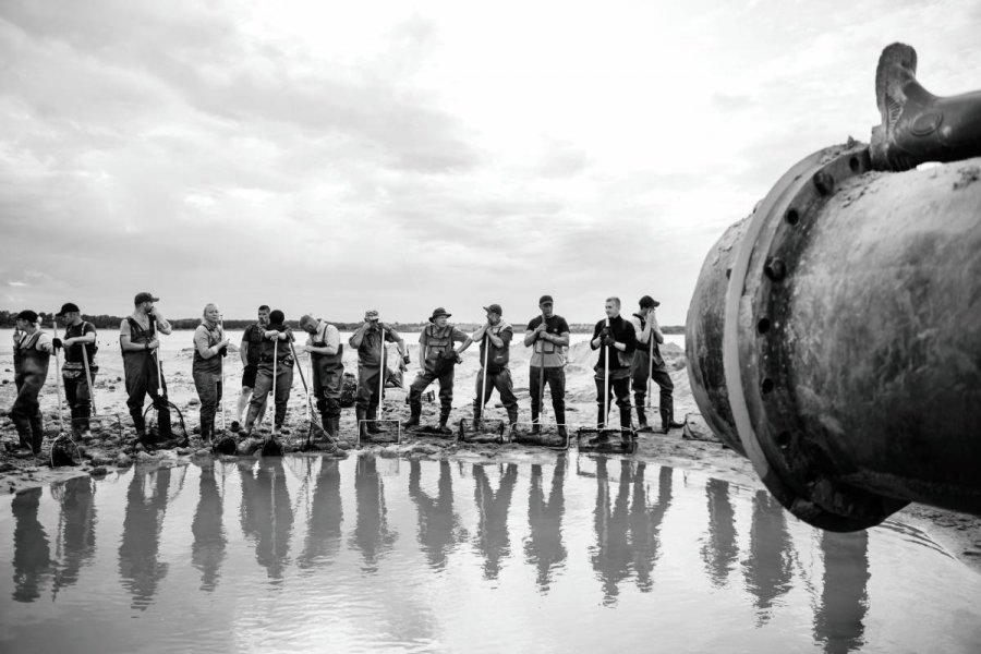 Czarno-biała fotografia przedstawiająca szereg mężczyzn z łopatami stojący przy dole wypełnionym wodą