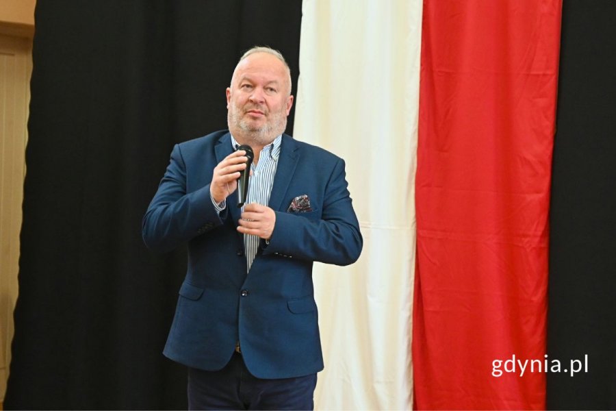 Mężczyzna stojący i mówiący do mikrofonu na tle granatowej dekoracji z motywem polskiej flagi