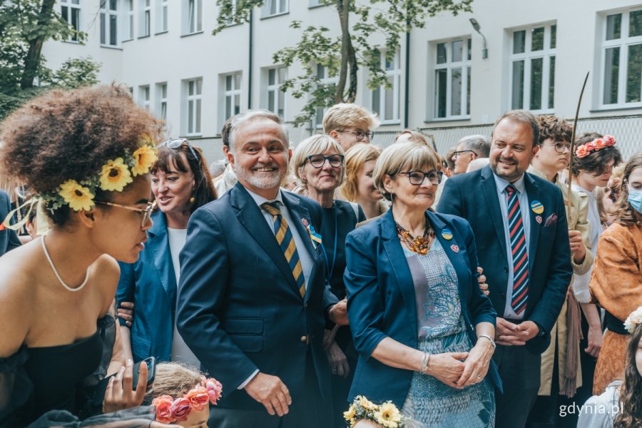 Grupa ludzi stoi na boisku szkolnym, smieją się.