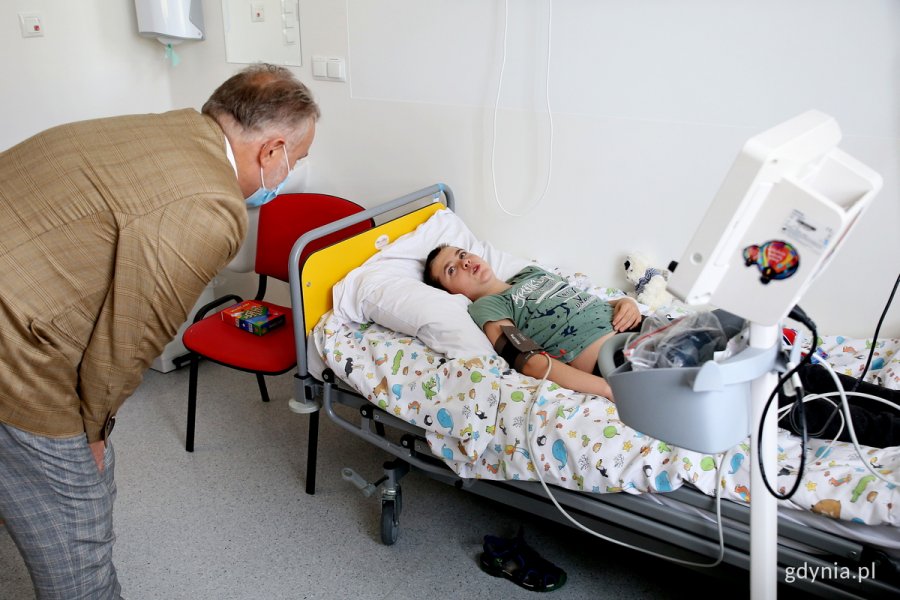 Prezydent Gdyni pochyla sie nad chłopcem leżącym na łóżku w pokoju szpitalnym.