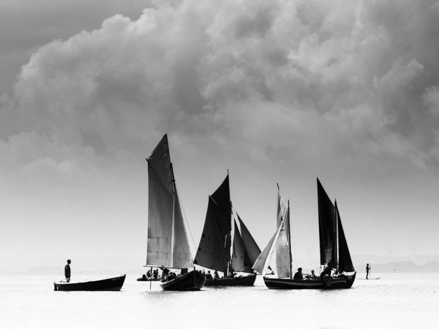 Czarno-białe zdjęcie przedstawiające kilka żaglówek i niewielką łódź na powierzchni morza