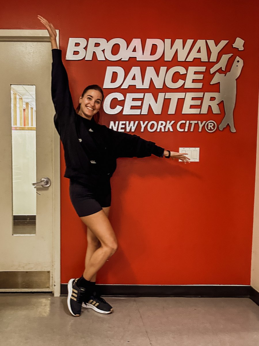 Uśmiechnięta tancerka na tle czerwonej ściany sali gimnastycznej z napisem Broadway Dance Center