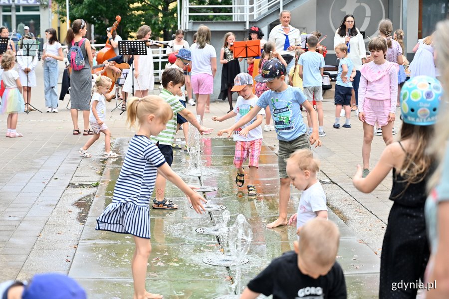 Grupa dzieci stoi przy fontannie na placu Kaszubskim - uczestniczą w spotkaniu z cyklu "Chodźcie na plac Kaszubski". W tle animatorki oraz uczniowie Szkoły Muzycznej I i II stopnia im. Z. Noskowskiego w Gdyni grający koncert // fot. Michał Puszczewicz