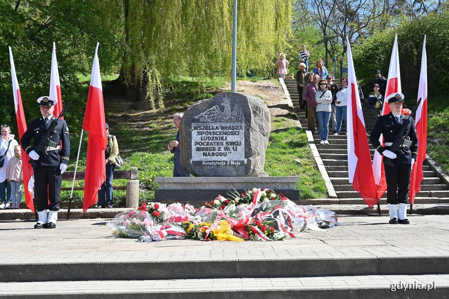 Obchody Narodowego Święta Konstytucji 3 Maja w Gdyni. Na środku zdjęcia: pomnik Konstytucji 3 Maja przy ul. Słowackiego i leżące przed nim wieńce oraz wiązanki kwiatów. Po bokach pomnika stoją marynarze, tworząc posterunek honorowy. Marynarze stoją na tle biało-czerwonych flag. W tle zieleń oraz mieszkańcy uczestniczący w uroczystościach // fot. Michał Puszczewicz