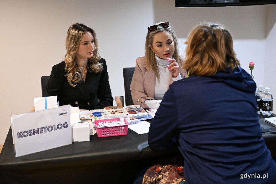 Trzy kobiety przy stoliku z tabliczką "kosmetolog", jedna z kobiet odwrócona plecami, pomieszczenie