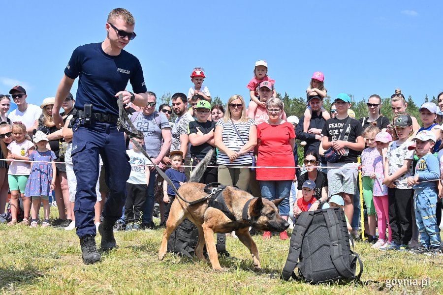 Policjant z wyszkolonym psem podczas pokazu na polanie. W tle stoją mieszkańcy