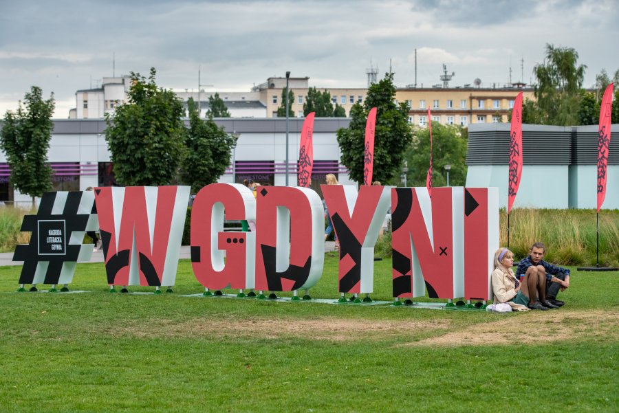Czerwono - czarny hashtag #wGdyni rozstawiony na trawie na placu Grunwaldzkim. Opiera się o niego dwoje ludzi. W tle czerwono-czarne flagi festiwalu "Miasto Słowa" // fot. Anna Rezulak