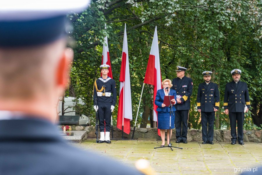 Barbara Pytko, prezes stowarzyszenia Gdyńska Rodzina Katyńska przywitała uczestników obchodów 81. rocznicy zbrodni katyńskiej. W tle: drzewa, biało-czerwone flagi i przedstawiciele Marynarki Wojennej// fot. Kamil Złoch