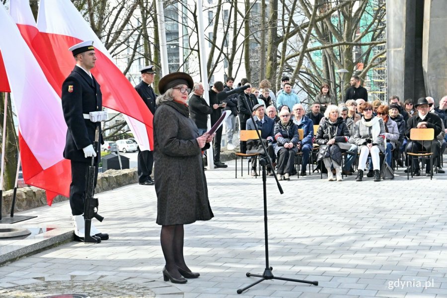 Barbara Pytko, prezes stowarzyszenia Gdyńska Rodzina Katyńska, przemawiająca podczas obchodów 83. rocznicy zbrodni katyńskiej, które odbyły się na Cmentarzu Wojennym w  Redłowie. Za nią marynarz na tle powiewających biało-czerwonych flag. W tle uczestnicy uroczystości // fot. Magdalena Czernek