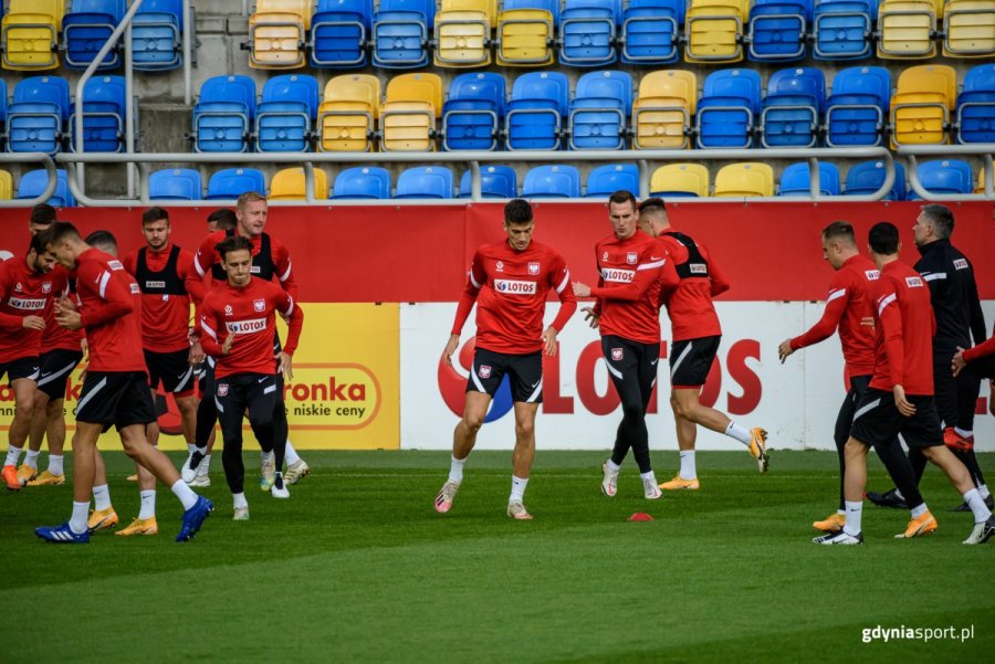 Zawodnicy w czerwonych bluzach i czarnych spodenkach podczas treningu na murawie stadionu. W tle żółto-niebieskie trybuny. 