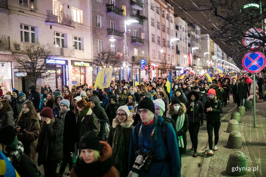 Mieszkańcy maszerują wieczorem ulicą podczas marszu antywojennego, trzymając flagi Ukrainy i hasła antywojenne.