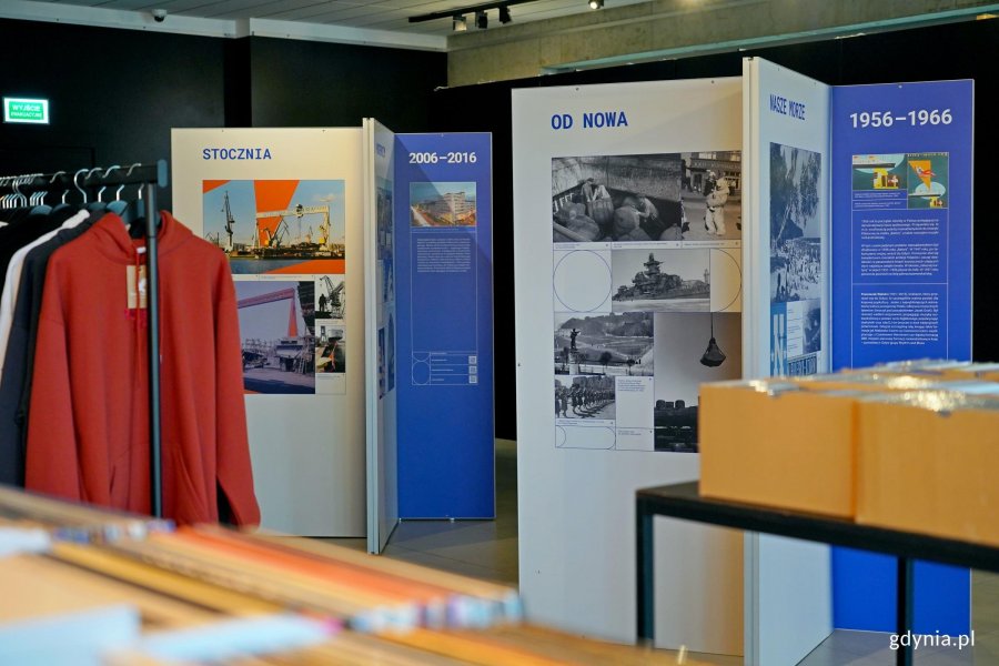 Ekspozytory z wystawą &bdquo;Gdynia x 10&rdquo; w Galerii Gdyńskiego Centrum Filmowego im. Leszka Kopcia stojące obok wieszak&oacute;w z bluzami i p&oacute;łek z płytami
