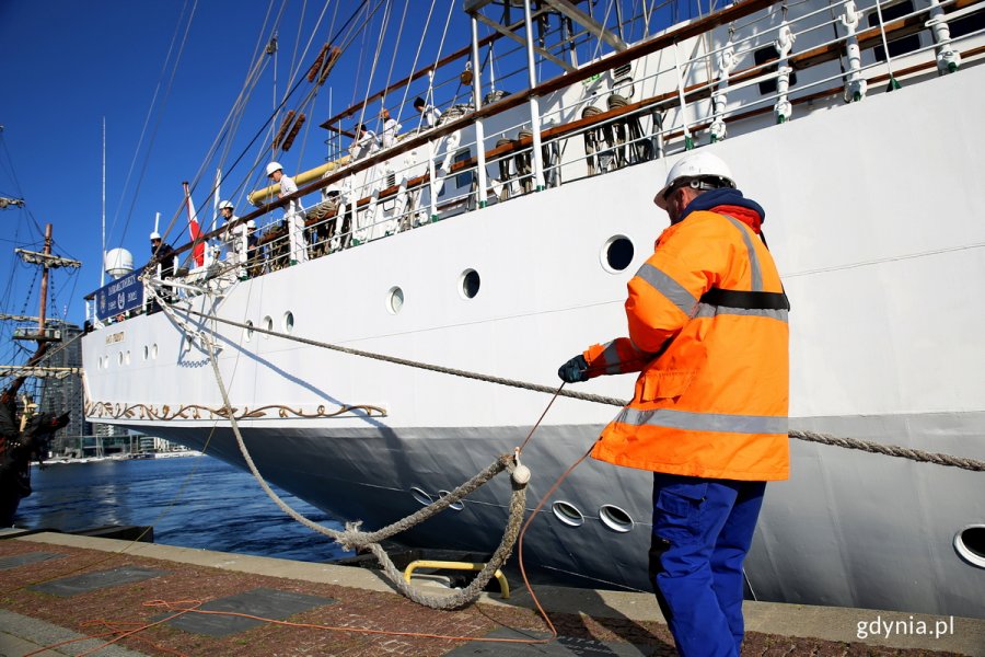 Na pierwszym planie mężczyzna w pomarańczowej kamizelce trzyma cumę „Daru Młodzieży”, który cumuje przy nabrzeżu Pomorskim // fot. Przemysław Kozłowski