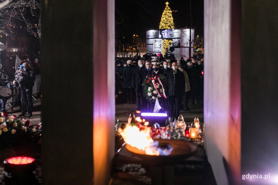 Na pierwszym planie płonąca pochodnia na pomniku Ofiar Grudnia '70 . W tle uczestnicy wieczornych uroczystości upamiętniających wydarzenia grudnia 1970 roku składający kwiaty przed pomnikiem Ofiar Grudnia '70. W oddali jest widoczna oświetlona lampkami choinka stojąca koło budynku Urzędu Miasta Gdyni // fot. Karol Stańczak