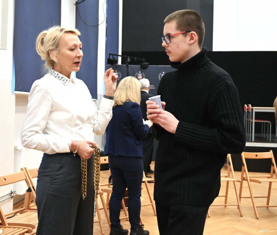 Kobieta w białej bluzce ze złotym łańcuchem z godłem i chłopak w czarnym golfie