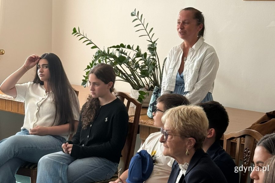 Przedstawiciele młodzieży z Gruzji podczas spotkania sali wizytowej, siedzący na fotelach. Za nimi kobieta-opiekun oraz stół i kwiat w doniczce.