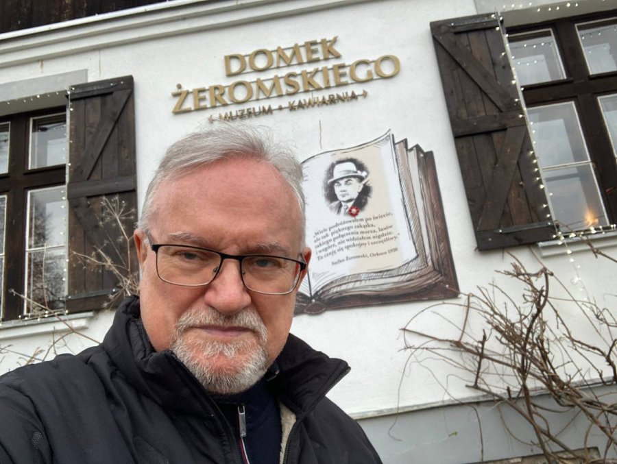Mężczyzna na tle białej ściany z oknami w brązowych okiennicach ozdobionej grafiką książki z twarzą Stefana Żeromskiego i szyldem z nazwą muzeum
