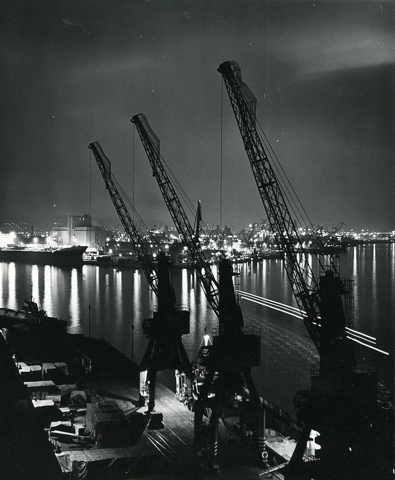 Fotografia przedstawia port w Gdyni po zmroku. Na pierwszym planie widok z góry na nabrzeże Fińskie z trzema, ustawionymi w rzędzie dźwigami portowymi. Na dalszym planie widoczny jest oświetlony port wewnętrzny, z lewej początek basenu im. Marsz. Józefa Piłsudskiego, w którym zacumowany jest statek handlowy. Widoczny przy nabrzeżu Indyjskim przed elewatorem zbożowym. Po prawej stronie, widoczne dźwigi portowe i światła portu w Gdynia. Fotografia pochodzi z archiwum cyfrowego Muzeum Miasta Gdyni // fot. Halina Wasielke