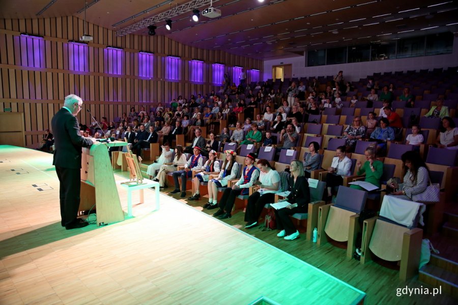 Ludzie zgromadzeni na sali konferencyjnej, jeden mężczyzna przemawia na scenie.