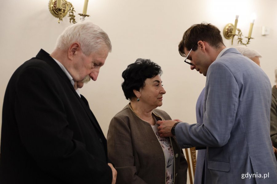 Mężczyzna w garniuturze przypina medal starszej pani w beżowej marynarce