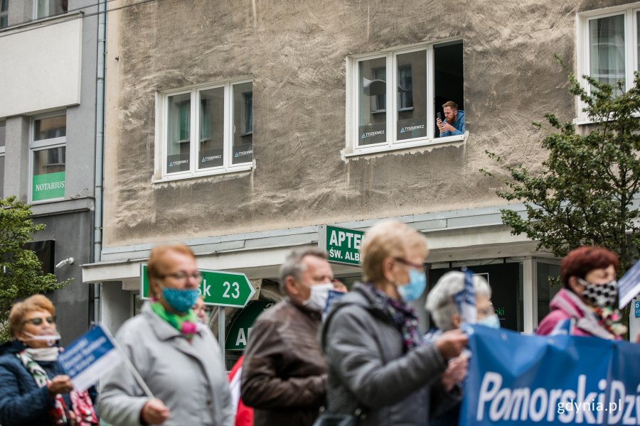 Wielobarwny Pomorski Przemarsz Seniorów ulicami Gdyni. W oknie kamienicy obserwujący ludzie. Fot. Karol Stańczak