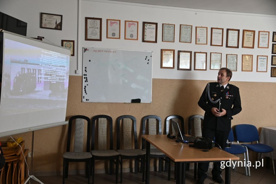Strażak w mundurze galowym stojący przy w sali przy ekranie i wygłaszający prezentację