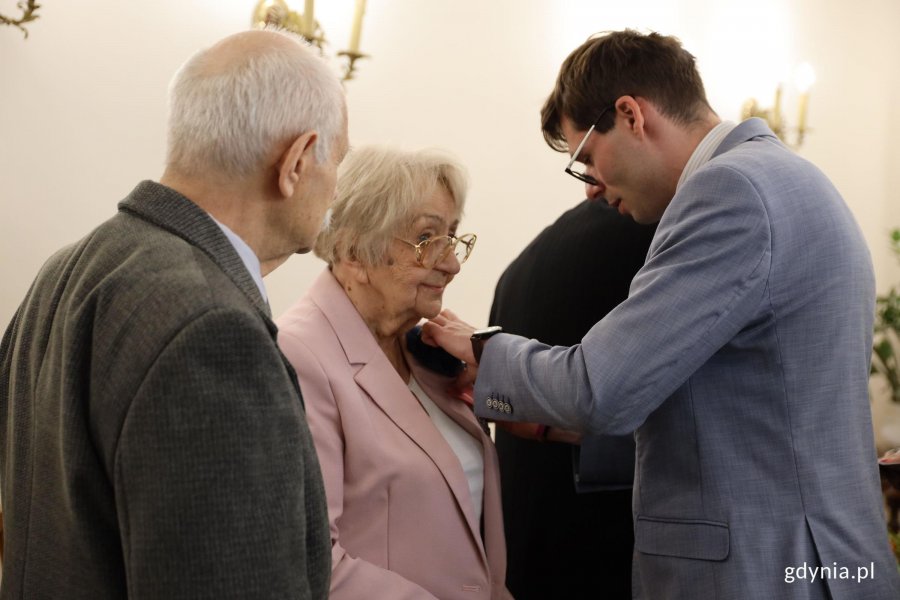 Mężczyzna w garniuturze przypina medal starszej pani w r&oacute;żowej marynarce
