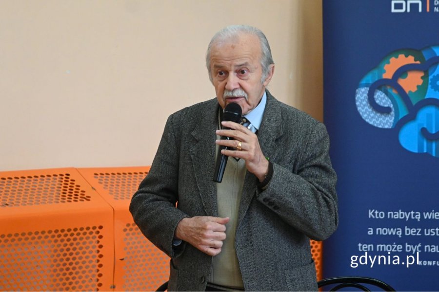 Senior stojący i mówiący do mikrofonu na tle beżowo-pomarańczowej ściany granatowej ścianki informacyjnej