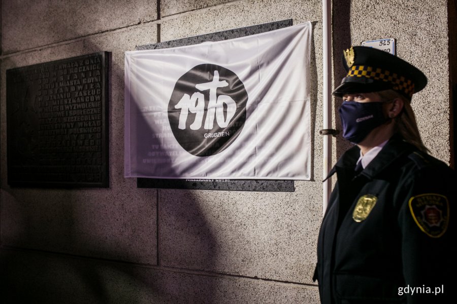Tablica pamiątkowa na fasadzie budynku Urzędu Miasta przed odsłonięciem - osłonięta białą flagą z czarnym znakiem Grudnia 1970. Przy tablicy stoi strażniczka miejska w mundurze // fot. Karol Stańczak