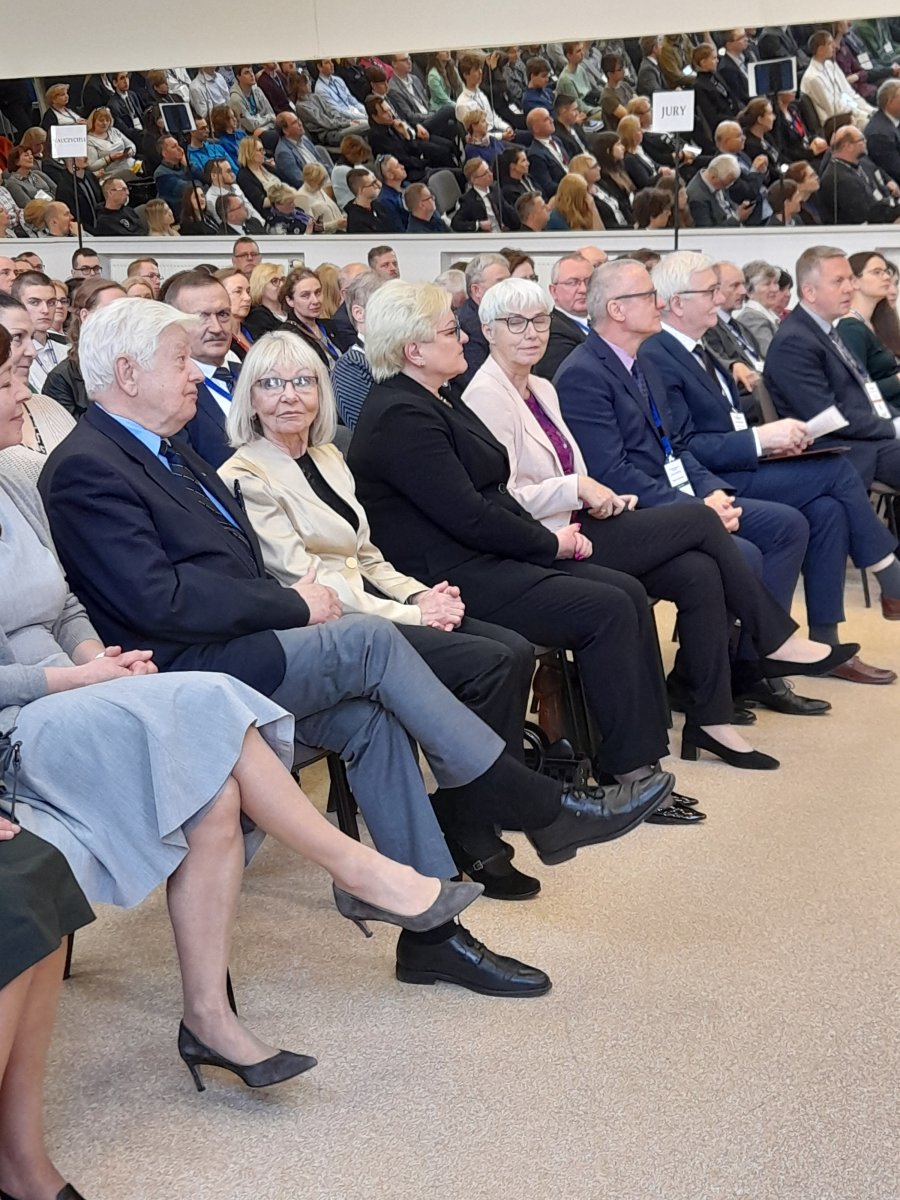 Na zdjęciu widać osoby w pierwszym rzędzie publiczności, w tle kolejne, które odbijają się też w lustarch zawieszonych wysoko na ścianie.