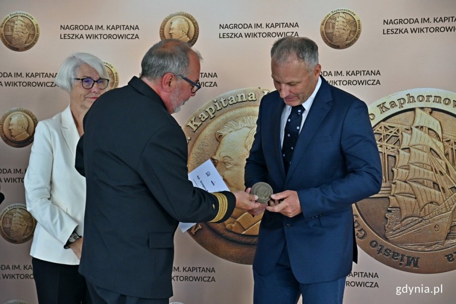 kpt. Grzegorz Baranowski odbiera nagrodę specjalną z rąk kpt. ż. w. Mariusza Wiktorowicza - syna patrona nagrody. Obok stoi Joanna Zielińska - członkini kapituły i radna Gdyni