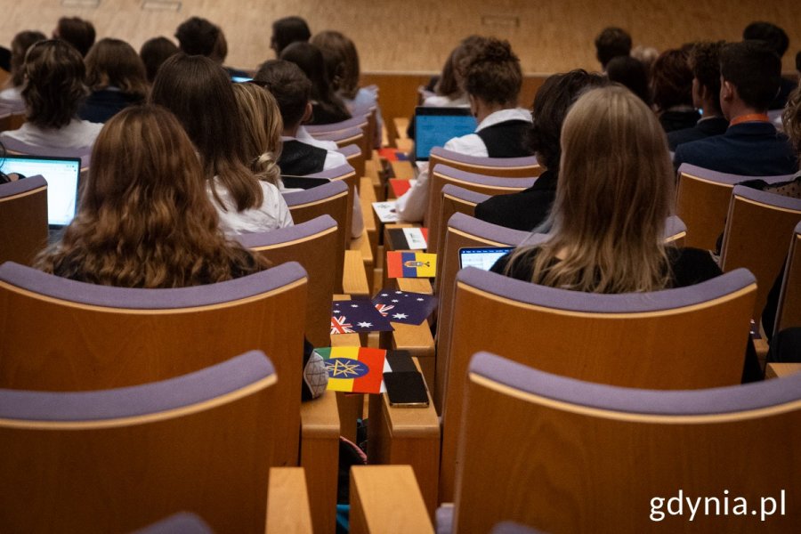 Uczestnicy konferencji siedzą tyłem, a między krzesłami są rozłożone flagi różnych państw